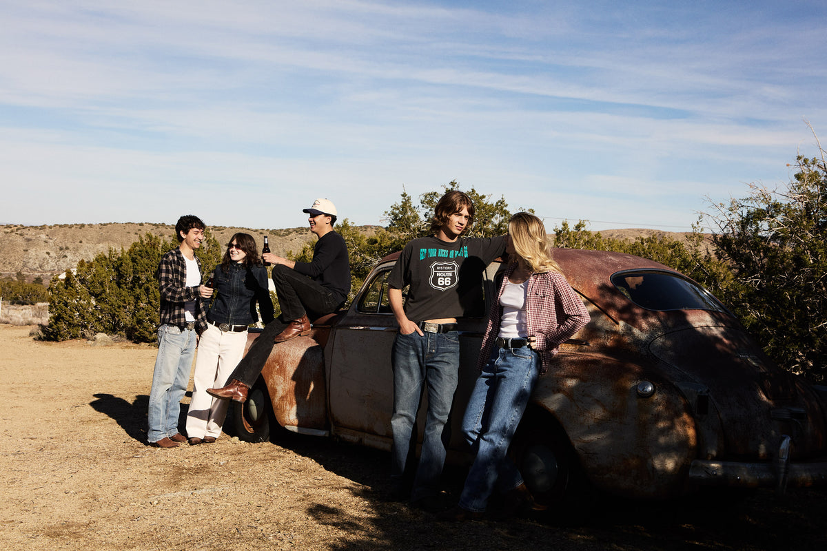 Group of people wearing belt buckles standing around a vintage car in a desert setting