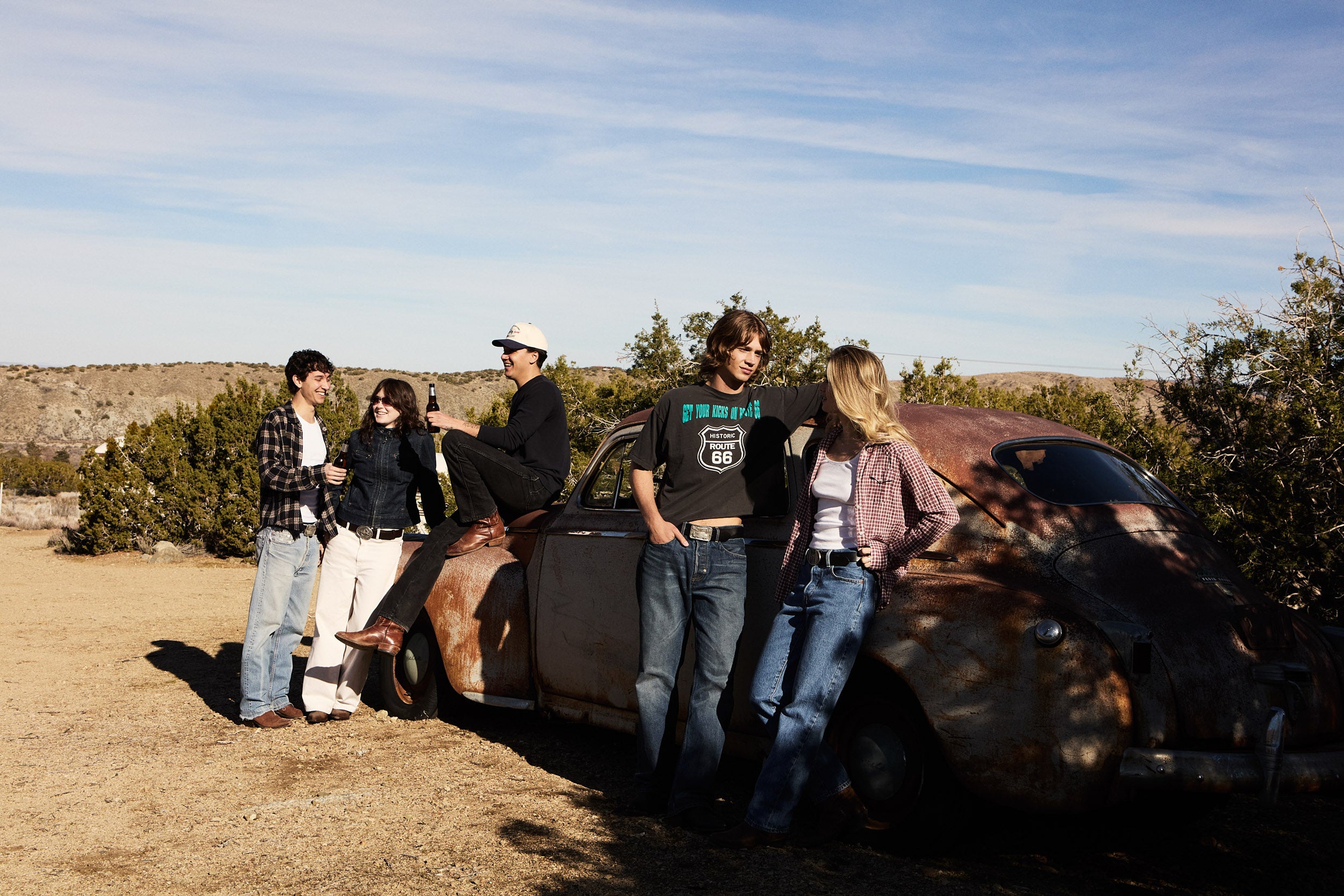 Group of people wearing belt buckles standing around a vintage car in a desert setting
