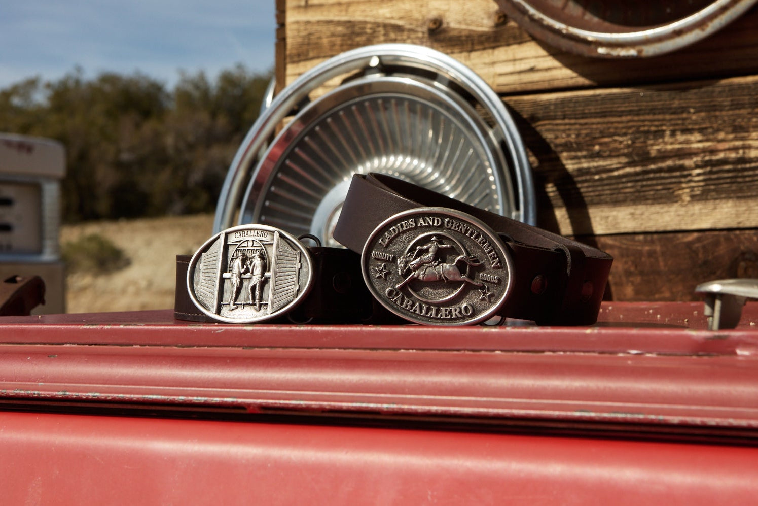 Two Caballero belt buckles on a red surface with a rustic background. The buckles are the Rodeo and Bar buckle